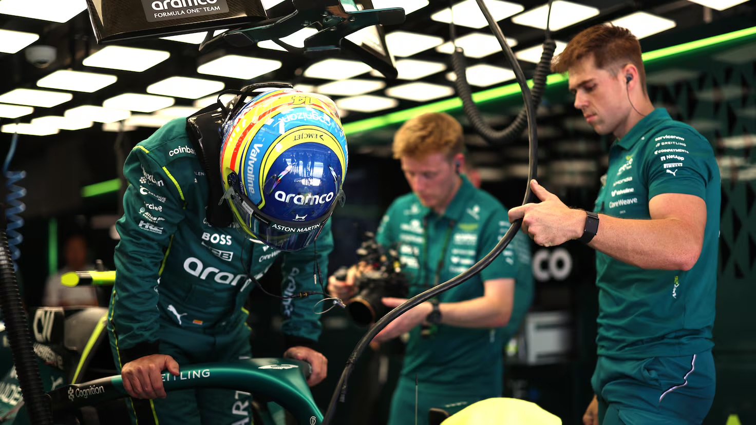Fernando Alonso se coloca el casco en el garaje de Aston Martin mientras ingenieros del equipo preparan el monoplaza
